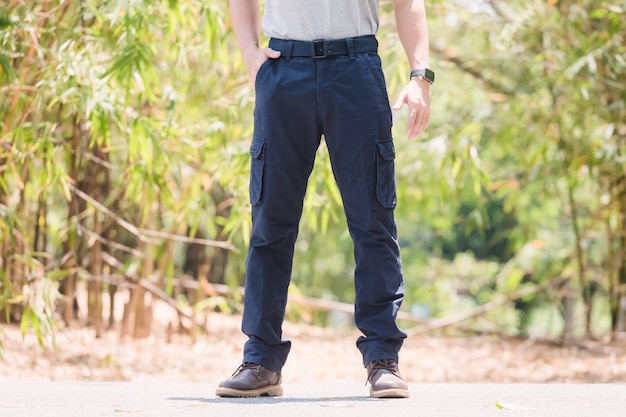 A person wearing dark blue cargo pants with multiple pockets, a light shirt, and brown shoes stands outdoors on a paved path surrounded by greenery.