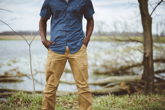 A person stands outdoors near a serene body of water, wearing a blue button-up shirt and mustard-colored pants.