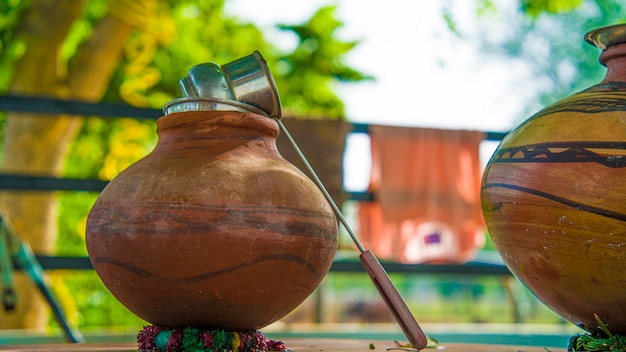 A traditional clay pot with a metallic lid and wooden ladle is displayed outdoors, accompanied by another colorful ceramic pot.