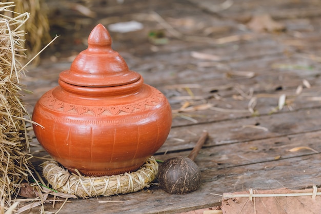 A rustic terracotta jar with a conical lid sits on a woven mat, surrounded by hay and a coconut.