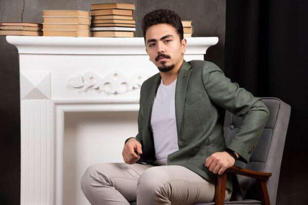 Man in a green blazer and beige pants sitting on a chair near a fireplace with books.