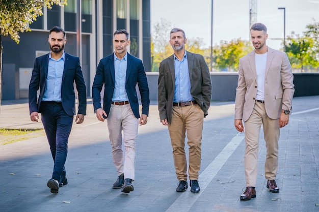 Group of four confident men in smart casual outfits walking outdoors, dressed in blazers, shirts, and trousers in neutral tones.