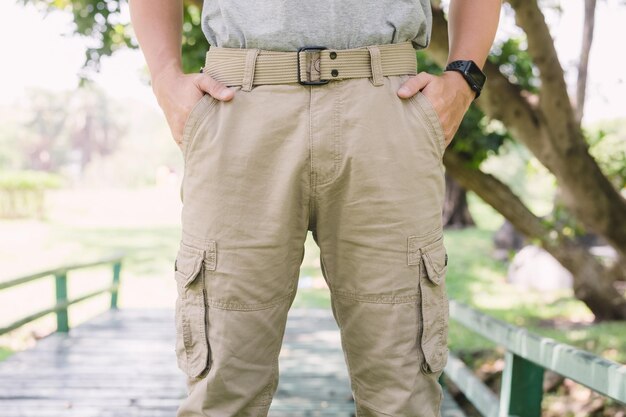Man wearing beige cargo pants with multiple pockets and a matching belt, standing outdoors on a wooden bridge.